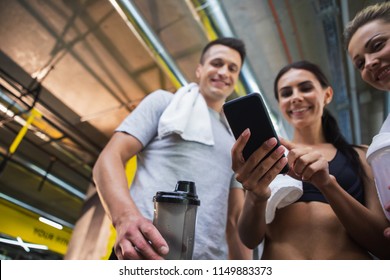 Low Angle Of Three Sporty People Using Mobile Phone In Gym With Focus On Gadget. Two Girls And Guy Are Standing And Looking At Device. They Are Holding Water Flasks And Towel