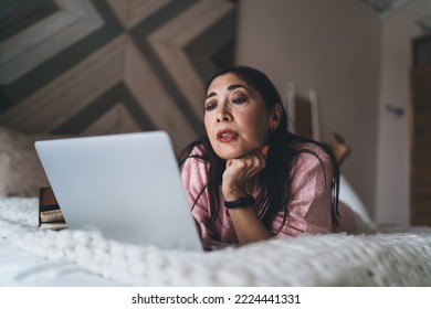 Low Angle Of Uncertain Middle Aged Asian Female Looking At Screen While Leaning Chin On Hand And Relaxing On Bed With Laptop And Watching Video In Free Time At Home. Black Friday Online Shopping