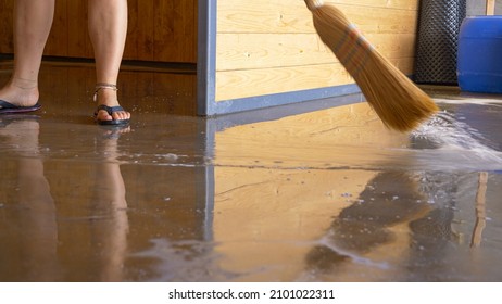 LOW ANGLE: Unrecognizable Woman Uses A Straw Broom To Sweep The Flooded Floor After A Monsoon. Young Female Wearing Flip Flops Sweeps Up Water Covering The Ground Floor Of Her House Under Construction