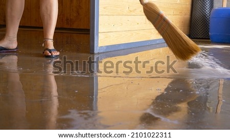 LOW ANGLE: Unrecognizable woman uses a straw broom to sweep the flooded floor after a monsoon. Young female wearing flip flops sweeps up water covering the ground floor of her house under construction