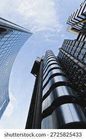 Low angle vertical shot of two distinct modern skyscrapers reaching into a light blue sky with thin white clouds The building on the left features a curved glass facade reflecting the sky and surrounding environment with a visible grid pattern of window panes The structure on the right side of the frame displays an industrial architectural style with prominent external silver metallic pipes and circular structural elements stacked vertically The metallic surfaces of the right building show light reflections and shadows that emphasize its complex mechanical design Both buildings are viewed from a ground perspective looking directly upward which creates a sense of scale and height The lighting is bright and natural highlighting the contrast between the smooth glass of the left skyscraper and the textured metal components of the right one This factual description captures the geometric lines and materials of contemporary urban architecture against a clear daytime sky
