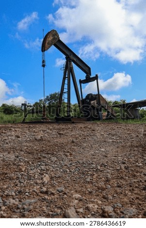 ​A low angle vertical view of an industrial oil pumpjack machine standing on a gravel ground under a blue sky with white clouds.