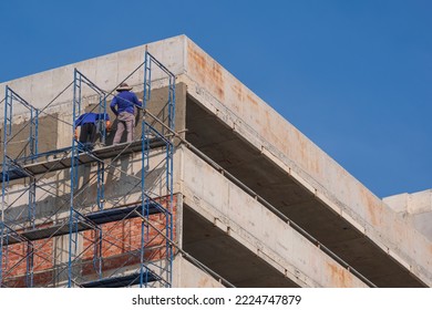 Low Angle View Of 2 Construction Workers On High Scaffolding Are Plastering Cement Wall On Rooftop Of Parking Garage Building In Construction Site Area