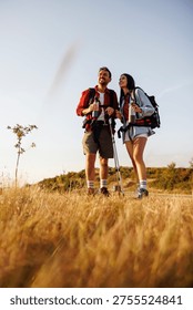 Low angle view of active couple carrying backpacks having fun hiking in nature on sunny summer day, taking a break relaxing and drinking water