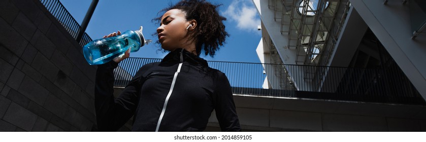 Low Angle View Of African American Sportswoman In Earphone Holding Sports Bottle On Urban Street, Banner