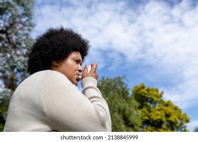 Low Angle View Of African American Mid Adult Woman Using Asthma Inhaler While Standing Against Sky. Unaltered, Healthcare, Asthmatic, Reliever Asthma Inhaler, Inhaling And Illness Concept.