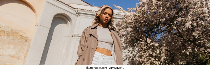 Low Angle View Of African American Woman In Stylish Outfit Standing Near Blooming Tree In Prague, Banner