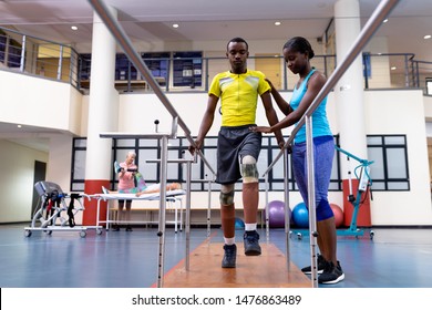 Low angle view of African-american physiotherapist helping disabled African-american man walk with parallel bars in sports center. Sports Rehab Centre with physiotherapists and patients working