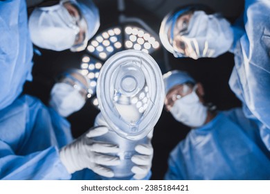 Low angle view of anesthetist holding oxygen mask above patient in surgery room with team of surgeon professionals. Operation on heart, brain, plastic surgery concept - Powered by Shutterstock