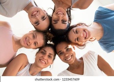 Low angle view of attractive diverse positive girls standing together in circle make huddle smiling looking from the top to camera. Women wearing sports tops ready to start workout at fitness studio