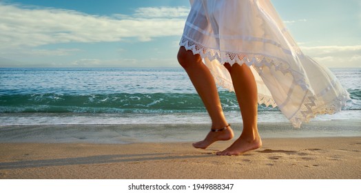 Low angle view of the bare legs of a woman in a flowing white skirt or dress walking along a sandy beach at sunset alongside gentle surf from a calm ocean