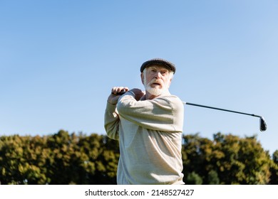 Low Angle View Of Bearded Senior Man In Flat Cap Holding Golf Club While Playing Outdoors