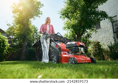 Low angle view of beautiful caucasian woman with blond hair using electric lawn mower for cutting grass on back yard. Pretty female in casual wear using modern equipment for work at garden.