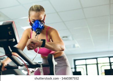 Low angle view of beautiful young Caucasian female athlete with oxygen mask checking time while exercising with exercise bike in fitness studio. Bright modern gym with fit healthy people working out - Powered by Shutterstock