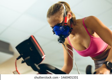 Low angle view of beautiful young Caucasian female athlete with oxygen mask exercising with exercise bike in fitness studio. Bright modern gym with fit healthy people working out and training - Powered by Shutterstock