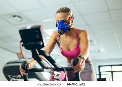 Low angle view of beautiful young Caucasian female athlete with oxygen mask exercising with exercise bike in fitness studio. Bright modern gym with fit healthy people working out and training - Powered by Shutterstock