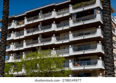 Low angle view between two palm tree trunks of a multi-story residential apartment building with curved white balconies and terraces under a blue sky