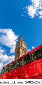 Low angle view of the Big Ben Clock Tower seen in great detail of its gold ornamentation and a red London double-decker bus passing below on a bright day with a clear blue sky and white clouds.