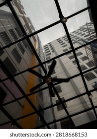 Low angle view of a black ceiling fan mounted under a glass skylight canopy, looking up at tall residential apartment buildings in Jakarta.