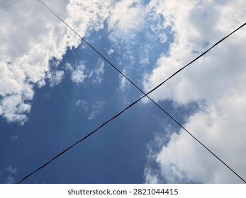 Low angle view of black electrical wires crossing forming an X shape against a bright blue sky with white clouds