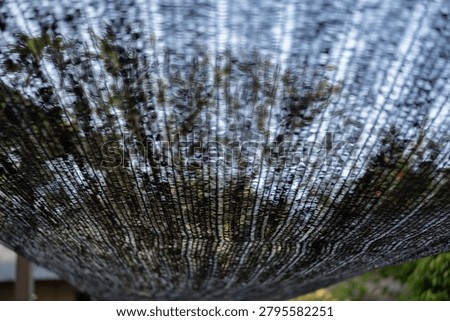 Low angle view of black plastic sun shade net with silhouettes of green trees blurred in the background