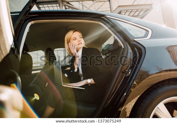 low angle view of blonde woman talking on\
smartphone while sitting with laptop in car\
