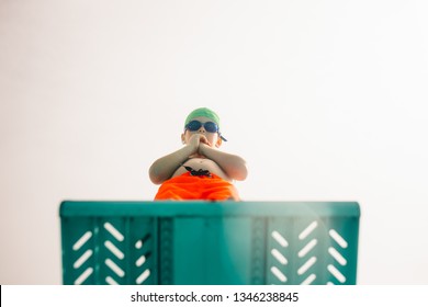 Low angle view of boy in swimming trunks, swim goggles and swimming cap standing on diving board against bright sky. Boy on diving platform at pool.