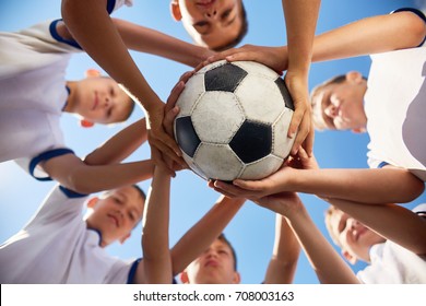 Low Angle View Of Boys In Junior Football Team Standing In Circle Holding Ball Together Against  Blue Sky, Focus On Ball