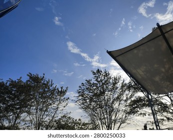 Low angle view of a bright blue sky with scattered clouds, framed by green trees and a modern outdoor shade canopy canopy structure. Sunny day background concept.