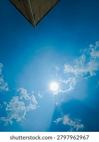 Low angle view of the bright sun shining through white clouds in a clear blue sky, with a sharp corner of a building roof at the top.