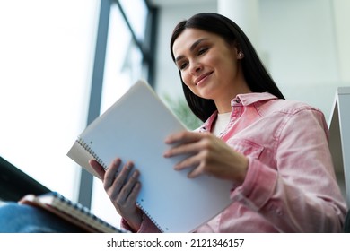 Low Angle View Of The Brunette Woman Student Sitting In The Library And Reading Book With Pleasure Smile. High School Student In The School Library Concept 