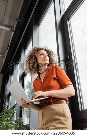 low angle view of businesswoman with curly hair holding laptop and looking through window in modern office
