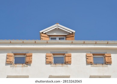 A low angle view captures a white plaster house facade featuring three small windows with open light brown wooden shutters and a dormer above against a clear blue sky