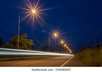 Low angle view of the car lights and starlight from roadside lamp posts from long exposure on a Thai rural road at dusk with cloudy morning and blue sky in the background.