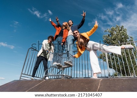 low angle view of cheerful interracial friends having fun with shopping cart on ramp in skate park