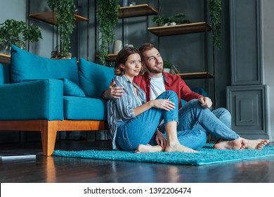 Low Angle View Of Cheerful Man Hugging Attractive Woman While Sitting On Carpet In Living Room 