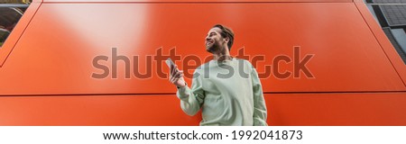 low angle view of cheerful man in sweatshirt holding smartphone near orange wall, banner