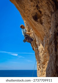 LOW ANGLE VIEW, CLOSE UP: Sport climber on an overhanging limestone route with the Mediterranean Sea on the horizon. Male athlete climbing against bright blue sky and sunlit rock in western Sicily.