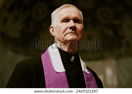 Low angle view closeup of elderly Caucasian Catholic preacher standing indoors looking at camera