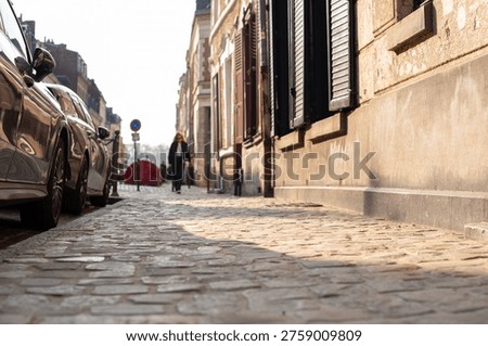 Low angle view of a cobblestone street and sidewalk in Lille, France. Historic brick and stone buildings with wooden shutters line the narrow road with parked cars.