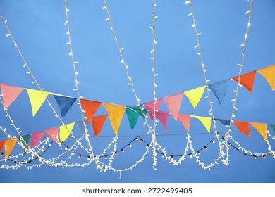 Low angle view of colorful triangular bunting flags and white string light garlands against blue evening sky. Colorful Holiday garlands