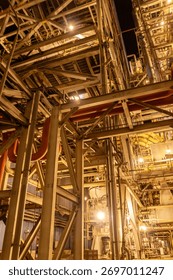 Low angle view of a complex industrial plant at night, featuring intricate metal structures, pipes, and warm lighting. Represents heavy industry, engineering, and infrastructure.