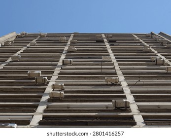 Low angle view of a concrete brutalist residential building facade covered with numerous air conditioning units against a clear blue sky.