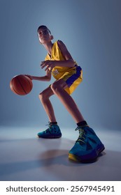 Low angle view of confident junior basketball athlete playing basketball in motion against grey studio background. Dribbling technique. Concept of sport, leisure activity, hobby, competition. Ad