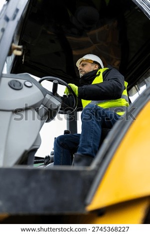 Low angle view of construction worker driving excavator at building site. Heavy machinery operator in protective helmet and reflective vest working on earthmoving project.