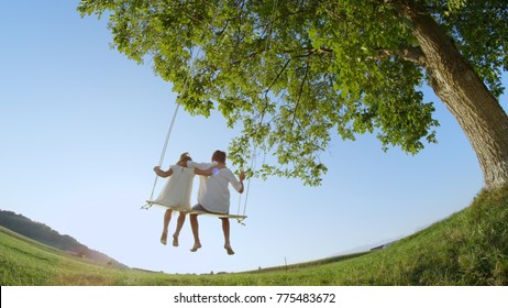 LOW ANGLE VIEW: Couple of young kids holding each other swinging on a wooden swing under a big green tree in summer. Boy and girl in embrace on a warm day. Hugging siblings swaying barefoot.