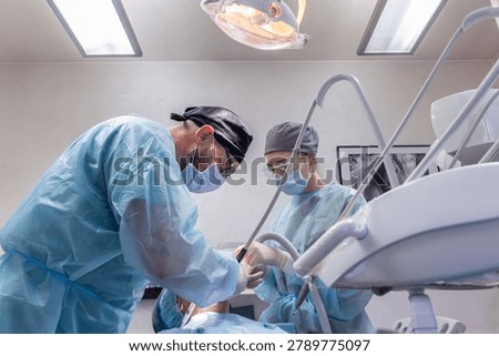 Low angle view of dentist and assistant performing oral surgery in a clinic, with overhead examination light protective gowns suction hose and covered dental unit framing focused teamwork around the patient