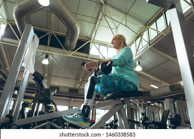 Low Angle View Of Disabled Active Senior Caucasian Woman With Leg Amputee Using Mobile Phone While Exercising In Fitness Studio. Strong Active Senior Female Amputee Training And Working Out