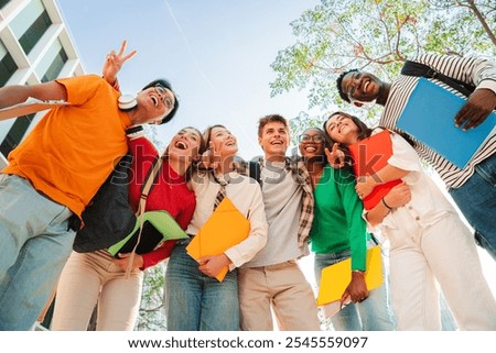 Low angle view of a diverse group of students smiling together, holding notebooks and backpacks, enjoying friendship and positivity outdoors, multicultural young friends bonding on campus. High