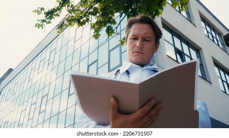 Low Angle View Of Doctor Looking In Folder With Documents Near Hospital Building